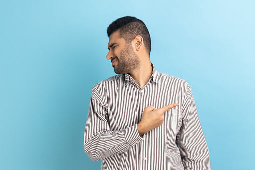 Get out. Portrait of serious angry businessman pointing finger, sneak male blaming another people, turning face, wearing striped shirt. Indoor studio shot isolated on blue background.