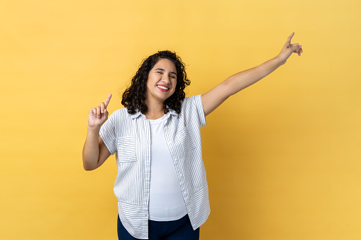 Portrait of extremely positive woman with dark wavy hair being in good festive mood, dancing, raising hands, expressing happiness. Indoor studio shot isolated on yellow background.