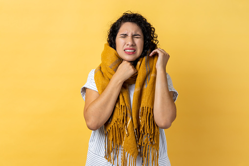 Portrait of sick woman with dark wavy hair in warm scarf, feeling unwell suffering fever, seasonal influenza symptoms and sore throat. Indoor studio shot isolated on yellow background.