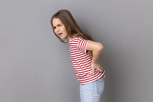 Side view portrait of little girl wearing striped T-shirt screaming in acute pain and holding sore back, risk of kidney stones disease, pinched nerve. Indoor studio shot isolated on gray background.