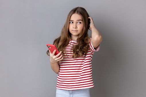 Portrait of pensive little girl wearing striped T-shirt using smart phone, looking away, thinking about post for her kid's blog. Indoor studio shot isolated on gray background.