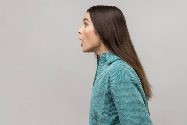 Side view of astonished woman with widely open mouth in amazement and big eyes looking ahead. Side view of astonished woman with widely open mouth in amazement and big eyes looking ahead, stunned shocked face, wearing casual style jacket. Indoor studio shot isolated on gray background. woman big eyes stock pictures, royalty-free photos & images