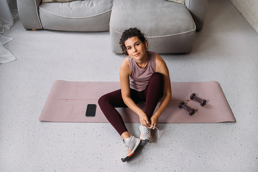 A happy young woman getting ready for a workout at home, sitting on a mat and tying her shoelace.