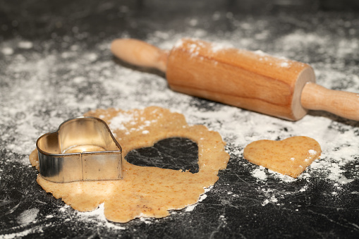 Cookie dough lies unrolled on a dark countertop. Flour is next to it and a rolling pin. A heart shape is on the dough, a heart has already been cut out.