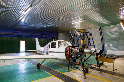 The hangar is a small business auto repair shop. Light single-engine aircraft, in need of engine repair. An aircraft engine is suspended on a chain hoist.