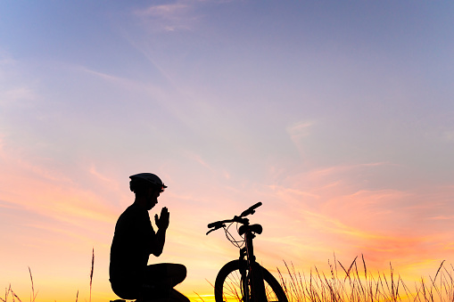 Silhouette of a man praying on the side of his mountain bike with the sunset sky in the background