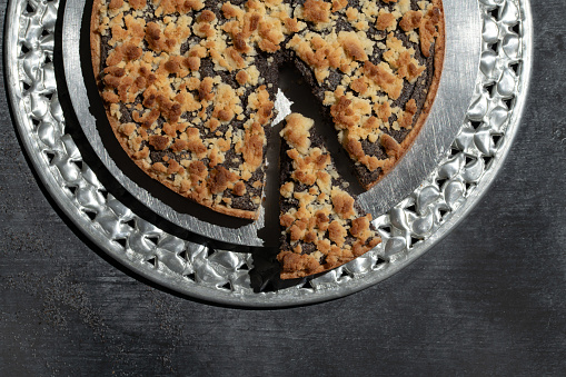 a freshly baked poppy seed cake with crumble is arranged on a cake plate. A piece is cut off and partially pulled out. The picture is photographed from above.