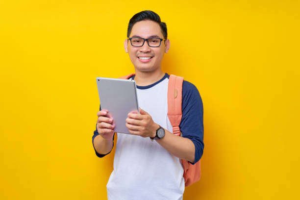 istock Cheerful young Asian man student in casual clothes glasses backpack using digital tablet computer for studying, reading e-books isolated on yellow background. high school university college concept 1529052188