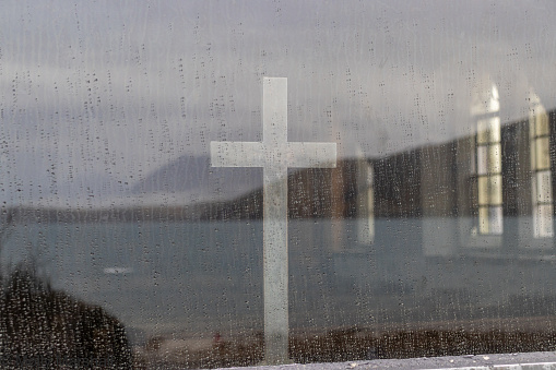 Close-up image of a cross inside a church viewed through a window from outdoors with rain on the glass.