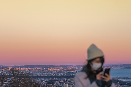 a solitary woman standing atop a hill, overlooking a sprawling cityscape. She is holding a phone in her hand and wearing a face mask, symbolizing the intersection of technology, personal safety, and urban life. The city below is a vibrant mosaic of lights and towering buildings, bustling with life yet observed from a distance. The scene exudes a sense of solitude and contemplation, as the woman takes a moment to connect with the city from her elevated vantage point. This photograph was taken at the Shonan coast in Enoshima, Kanagawa Prefecture, Japan.