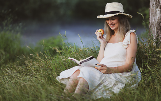 Young pregnant woman sits on a grass in the park with the book.