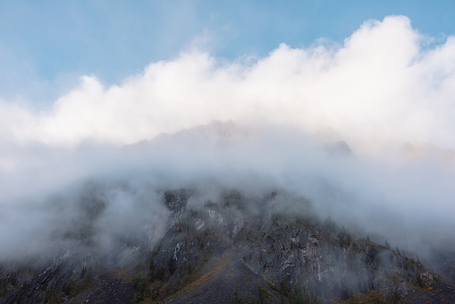 Lovely scenery with big mountain peak in thick clouds at early morning. Scenic view to high pinnacle in gantly blue cloudy sky in sunlight. Large mountain top in low clouds. Sharp rocks in dense fog.
