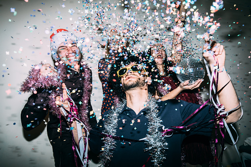 Four young adults are enjoying a party, wearing santa hats, tinsel and sunglasses and throwing confetti in the air