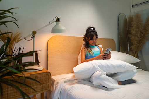 Young Asian woman sitting on the bed in bedroom and using mobile phone for online text message or social media at night. Attractive girl enjoy indoor lifestyle with using wireless technology at home.