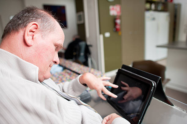 Mature aged man with a disability operating touchscreen computer Mature aged man with a disability operating touchscreen computer.  The reflection of his hand can be seen on the screen.  He is seated in his living room. developmental disability stock pictures, royalty-free photos & images