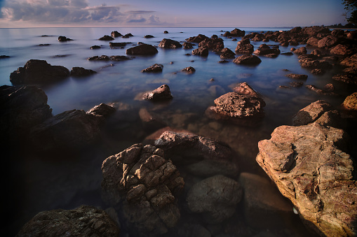 In the serene morning glow, a captivating photograph portrays a tranquil rocky beach with the calm sea glistening in golden hues, creating a picture-perfect moment of serene beauty.
