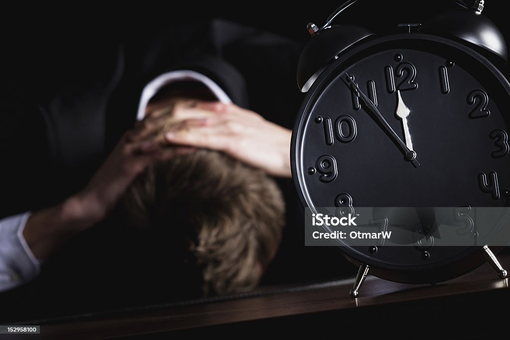 Businessman totally desperate. Desperate business person in dark suit sitting at office desk with head down being in despair with close up of alarm clock in foreground showing five minutes to twelve o-c'clock, low-key style isolated on black background. 30-39 Years Stock Photo