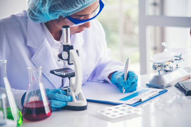 istock Woman scientist hand write note information in medical lab look at science microscope medical test, research biology chemistry. Asian Females technician laboratory analyzing blood test pharmaceutical 1529590796