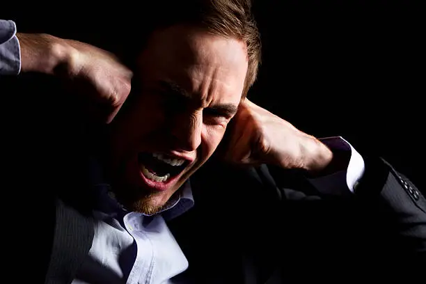 Low-key portrait of desperate businessman in dark suit screaming and holding both fists at head expressing strong despair, isolated on black background.