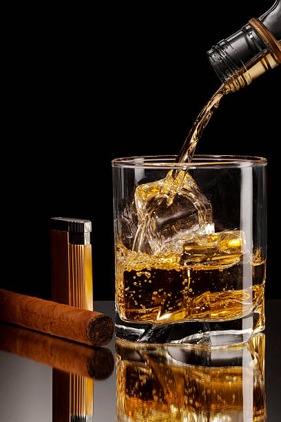 Glass of alcohol on the rocks shot on a reflective surface. Vertical shot with copy space. Cigar and Lighter beside. Alcohol being poured into the glass