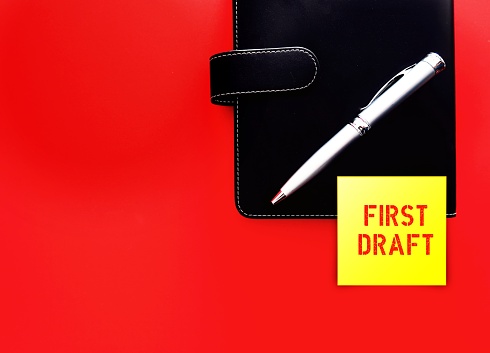 Black notebook and pen on red background with  stick note written FIRST DRAFT, concept of writing process, very first version of a piece of writing