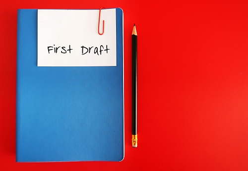A note written FIRST DRAFT clip with a blue notebook, with a pencil, isolated on red background. concept of writing process, very first version of a piece of writing, book or story