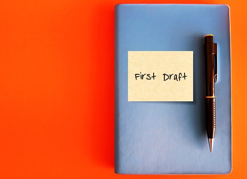 A blue notebook with stick note written FIRST DRAFT and a black pen isolated on orange background- concept of writing process, very first version of a piece of writing, book or story