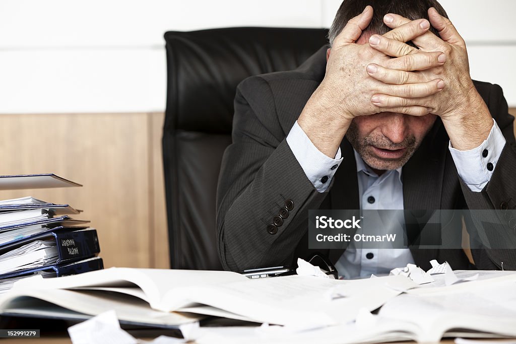 Frustrated businessman at desk with head in hands Worried businessman sitting at office desk full with books and papers being overloaded with work. Mental Burnout Stock Photo