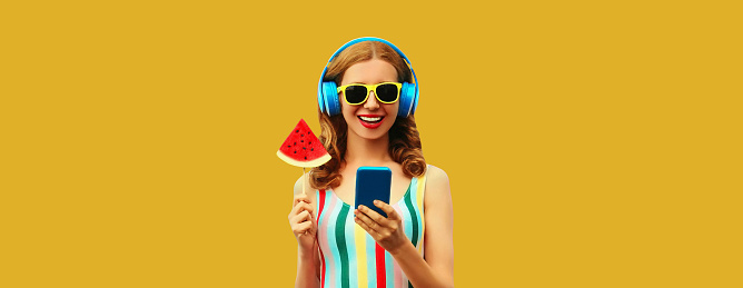 Summer portrait of happy smiling young woman model in headphones listening to music on smartphone with juicy lollipop or ice cream shaped slice of watermelon on yellow background