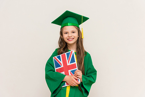 A student in a master's suit holds a book with the flag of Britain in English and smiles happily.  Language, educational school for children. Graduation in an English school. White isolated background