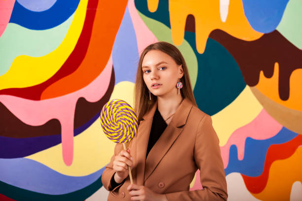Portrait of young woman holding round giant lollipop at colored wall, looking at camera. Pretty lady in beige suit posing with candy on stick. Summer fashion style concept. Copy ad text space, poster retrato de una mujer joven sosteniendo una piruleta gigante redonda en una pared de colores, mirando a la cámara. bonita dama con traje beige posando con caramelos en el palo. concepto de estilo de moda de verano. copiar espacio de texto del anuncio, pós