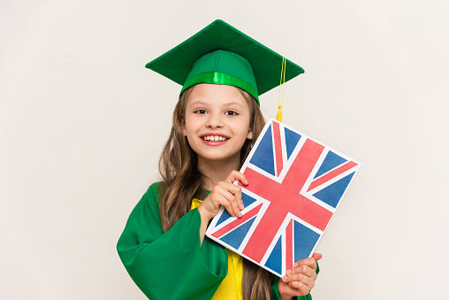 A student in a master's suit holds a book with the flag of Britain in English and smiles happily.  Language, educational school for children. Graduation in an English school. White isolated background