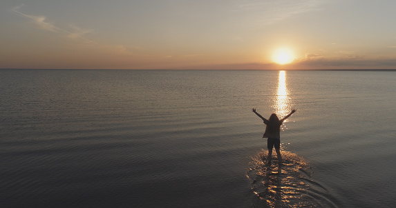 Aerial shot over teen girl barefoot running and raising arms in water on baltic sea beach in sunset time, wide photo