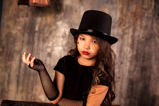 Portrait of child asian girl in black stylish image with hat posing in dark old room at textured wall background, looking at camera. Kid in fashionable costume. Theatre performance concept. Copy space