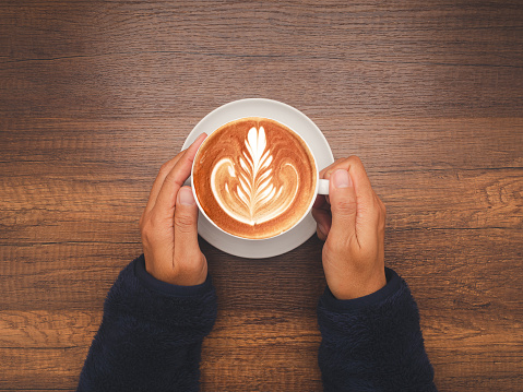 Top view of hands woman in sweater holding a fresh coffee cup over wooden table. Space for text. Beverage and relaxation concept