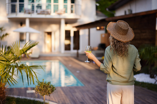 Rear view of a woman with hat holding glass of lemonade while standing by the pool in the backyard of her house.