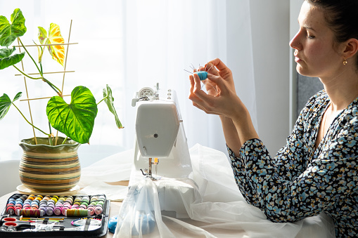 A woman sews tulle on an electric sewing machine in a white modern interior of a house with large windows, house plants. Comfort in the house, a housewife's hobby