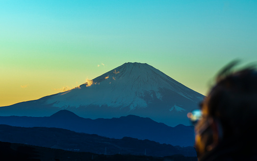This captivating image captures a tranquil scene at sunset, with the majestic Mount Fuji in Japan as its centerpiece. The mountain’s imposing silhouette dominates the upper half of the composition, beautifully outlined against a vibrant sky painted in hues of orange, purple, and gold. In the foreground, a lone figure stands in awe, his silhouette harmoniously blending with the serene surroundings. The fading sunlight casts a warm glow upon the rugged terrain, creating an enchanting contrast against the darkening sky. This image encapsulates the awe-inspiring beauty of nature, transporting the viewer to a serene moment where the majesty of the mountains and the tranquility of the sunset converge in sublime harmony.
