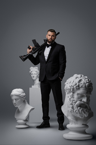Stylish man with a well-groomed beard dressed in a luxurious black suit and bow tie, holding an automatic rifle striking a confident pose among three antique statues against a grey background