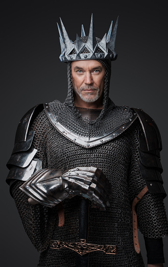 Medieval-style portrait of a charismatic king with a gray beard in chainmail armor with steel plates, holding a sword and posed against gray