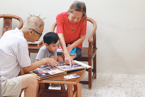 An Asian grandparents and their grandson sit in the living room, engrossed in conversation while looking through a photo album. They bond and cherish precious family moments, spending quality time together.