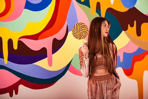 Lovely teen girl with round giant lollipop posing at colored wall, looking away. Portrait of teenage cover girl model in stylish beige wear with candy on stick. Sweet life concept. Copy ad text space