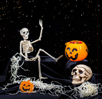 A toy Skeleton crawling out of the grave, and waving his hand, greets, pumpkins with grimaces, a skull on a black background. The eve of the day of death