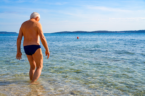 Senior men a Parkinson Disease patient carefully entering the sea water. He is trying to cool down before entering the water. It is early spring near Peroi, Istrian coast in Croatia.