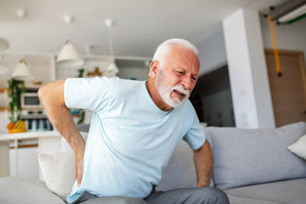älterer mann mit rückenschmerzen berührt seinen rücken und veranschaulicht ischias und sitzende lebensweise. betonung der wirbelsäulengesundheit und der bedeutung von gesundheitsversorgung und versicherung in diesem stockbild. - arthritis senior adult rheumatoid arthritis sadness stock-fotos und bilder