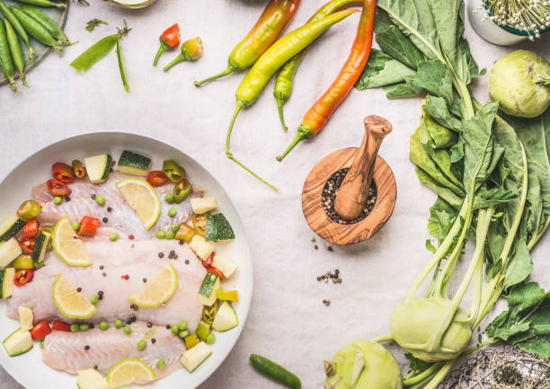 Keto dietary fish dish with vegetables in white pan on light kitchen table background with ingredients and knife, top view. Cooking preparation. Healthy low carb meals concept. Diet food concept