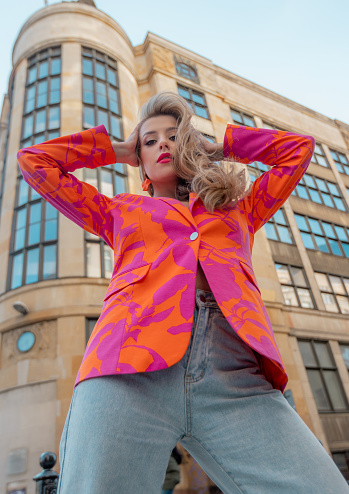 Elegant young lady with pink lips in colorful blazer standing on background of high-rise building in city. Confident woman with orange earrings raised hands and straightening blond hair. Vertical.