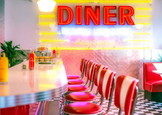 A retro styled diner with an old-fashioned glowing neon sign, and a row of seats arranged by the counter.