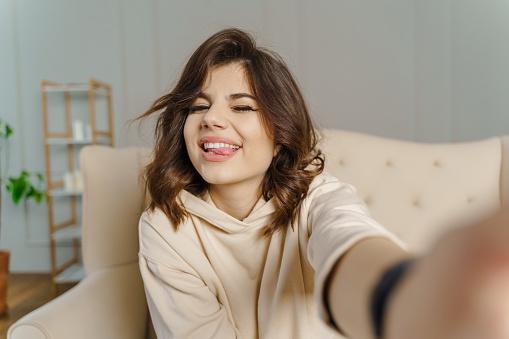 Pretty young asian female with big smile sitting at living room. She having fun taking light cheerful selfie on blurred background. High quality photo