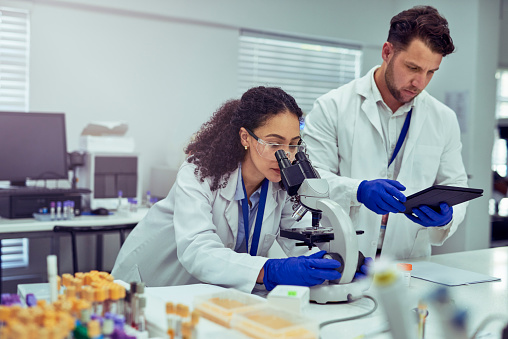 Female scientist wearing safety goggles and gloves with male colleague working together in laboratory, looking at test sample under microscope and using digital tablet